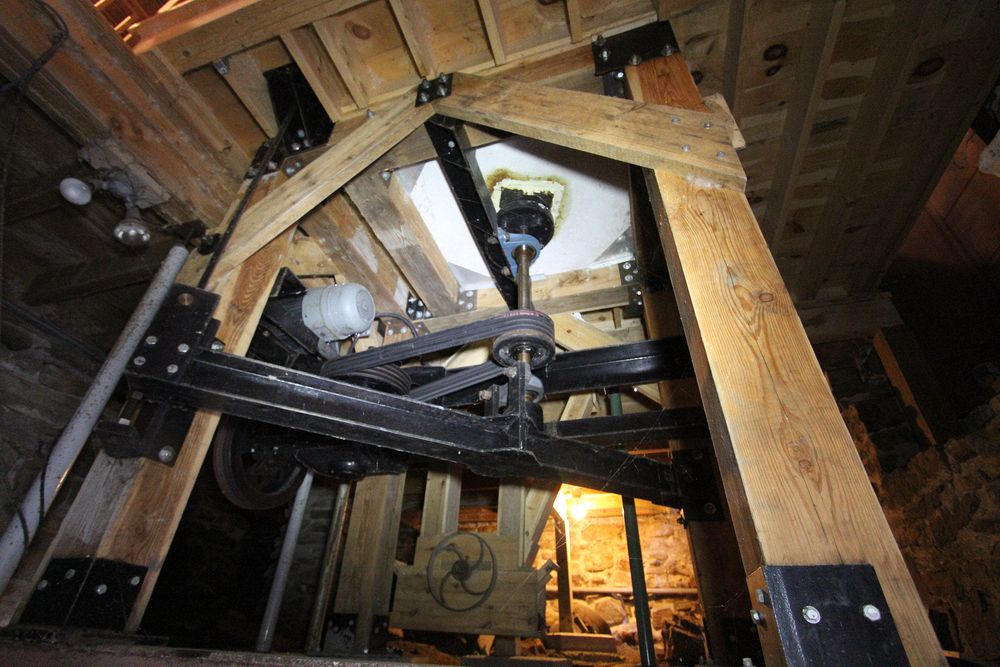 The centre shaft goes through the bedstone up to the runner (top) stone.  It is only the top stone that rotates, today at about 92 rpm.  Originally the stones turned faster, likely at about 98 rpm to speed up production.  It also heated the flour, the first stop for the flour was a "hopper boy" on the third floor that stirred the flour to help cool it and ensure that it didn't stick together.\n\nIn the background (metal wheel on wooden structure) is the "elevator boot" where the flour from the millstones is picked up  by the elevator (a continuous belt with small metal buckets) and taken to the third floor.